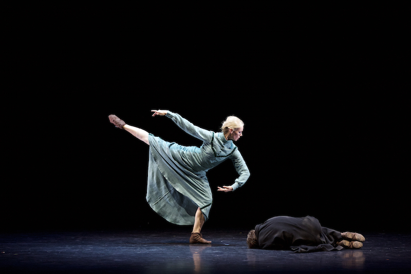 A woman in a long green dress plies on one leg peering over a man crumpled on the floor. Her other leg extends high in the air.